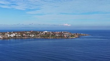 Gün doğumunda yumuşak mavi gökyüzüyle sakin bir sahil manzarası. Renkli evler kıyı şeridini işaretliyor, çevresindeki doğal manzara ve sakin sularla uyumu yansıtıyor..