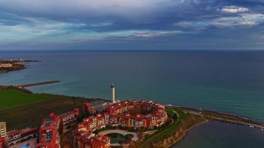 Resmedilmeye değer bir kıyı şeridine bakan sahne, hareketli sulara karşı kurulmuş, karamsar bulutlar ve sakin bir ortam ile tamamlanmış zarif bir kıyı mimarisini yakalıyor..