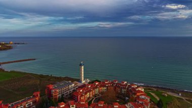 Büyüleyici bir kıyı köyü kırmızı çatılı binalar, yükselen bir deniz feneri rehberlik eden tekneler ve alacakaranlık renklerini yansıtan sakin bir deniz, bulutlar gökyüzünde sürüklenirken.