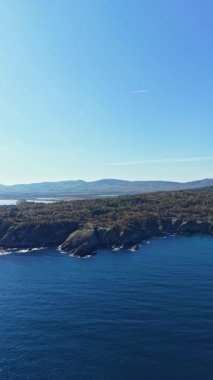 Dalgalar, güneş suyun üzerinde dans ederken engebeli taşlara nazikçe çarpar. Uzak tepeler parlak mavi gökyüzüne karşı huzurlu bir zemin oluşturuyor. Doğanın huzur verici güzelliğini gözler önüne seriyor..