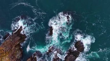 Kıyı şeridi boyunca uzanan karanlık, pürüzlü kayalara çarptıklarında güneş ışığı dalgaların üzerinde dans eder. Turkuaz su kayar ve kıvrılır, doğanın gücünün büyüleyici bir sahnesini yaratır..