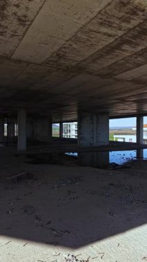 Boş bir yapının içinde, güneş ışığı büyük açıklıklardan geçerek çatlamış betonun üzerine gölgeler düşürür. Yerdeki su birikintileri molozların arasında huzurlu yansımalar yaratıyor..