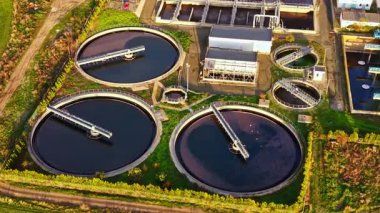 Bir su arıtma tesisinde parlak bir gökyüzünün altında suyu işleyen büyük dairesel tanklar bulunur. Bereketli yeşillik endüstriyel yapıları çevreleyerek, çevre dostu bir atmosferi vurguluyor.