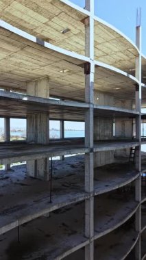 İşçiler, açık mavi gökyüzü altında, doğa ve kentsel gelişim arasındaki zıtlığı somutlaştıran, açığa çıkmış beton seviyelerini ve kirişleri gösteren, suyun yakınındaki eksik bir yapıda ilerliyorlar..