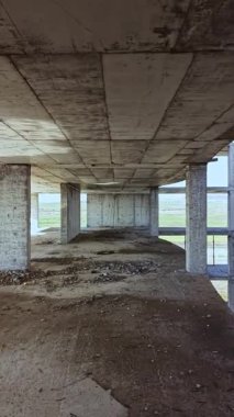 Güneş ışığı, kirli zeminleri ve pürüzlü dokuları gözler önüne sererek iskelet yapısının açık beton duvarlarından geçerken, yemyeşil alanlar arka planda ufuk boyunca uzanır..