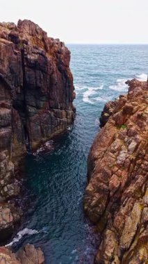 Dalgalar sakin bir sahil sahnesinde kadim kaya oluşumlarına hafifçe çarpar. Dalgalı mavi su kayalıkların toprak tonlarıyla çelişiyor ve güzel bir ahenk oluşturuyor..