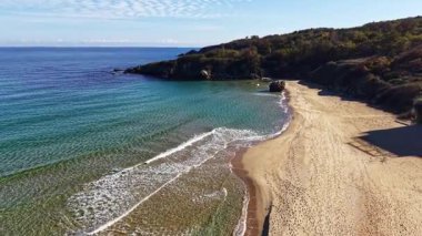 Yumuşak dalgalar altın kuma vurur. Güneş, kayalık kayalıklar ve canlı yeşilliklerle çevrili sakin bir plajda ışıldar. Huzurlu bir atmosfer yaratır..