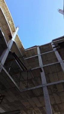 Terkedilmiş bir yapının içinden yukarıya doğru bakan berrak mavi gökyüzü, ham beton kirişler ve kısmi duvarlarla çelişiyor. Ürkütücü ama büyüleyici bir kentsel bozulma görüntüsü yakalıyor..