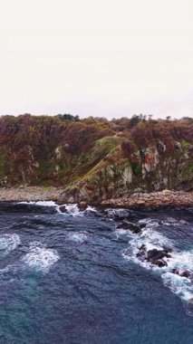Heybetli dalgalar, dalgalı yeşilliğin taşlı kıyı şeridiyle buluştuğu engebeli kayalıklara nazikçe çarpıyor. Huzurlu sahne, kıyı sükunetinin özünü yakalıyor..