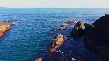 Sakin okyanus dalgaları, kıyı boyunca uzanan karanlık ve pürüzsüz kayalara doğru yavaşça suyla huzurlu bir öğleden sonra, parlak güneş ışığıyla aydınlanır..