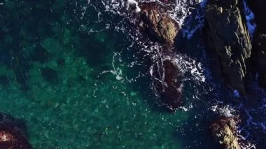 Turkuaz sular engebeli kayaların etrafında kıvrılır, dalgalar kıyıya vurur. Parlak güneş ışığı, canlı sualtı yaşamını aydınlatır. Huzurlu bir sahil manzarası yaratır..