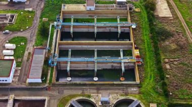 Güneşli bir günde açık mavi bir gökyüzünün altında yemyeşil çimlerle çevrili büyük dikdörtgen havuzlar ve boruların bulunduğu bir su arıtma tesisi..