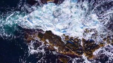 Parlak güneş ışığı turkuaz sularda parlarken güçlü dalgalar karanlık ve engebeli kayalara çarpıyor ve sahil şeridinde doğanın gücü ve güzelliğinin çarpıcı bir gösterisini yaratıyor..