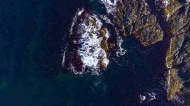 Güçlü dalgalar sert kayalara çarparak sakin mavi sularda beyaz köpük oluşturur. Güneş arka planda batıyor, sıcak renklerle gökyüzünü boyuyor..