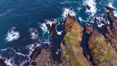 Bu çarpıcı sahil sahnesinde, güçlü dalgalar pürüzlü kayalarla çarpışır ve berrak mavi gökyüzünün altında beyaz su püskürtür. Canlı yeşil bölgeler manzaraya hayat katıyor..