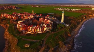 Manzaralı bir sahil köyünde canlı binalar ve uzun bir deniz feneri bulunur. Güneş yeşil alanlar üzerinde batar, aşağıdaki okyanus dalgalarına sıcak renkler dökerek sakin bir atmosfer yaratır..
