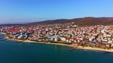 Sahil kasabası kıyı şeridi boyunca uzanır, manzarayı noktalayan canlı evlerle, sakin dalgalar parlak güneşli bir gökyüzünün altındaki kumlu sahilde tur atar..