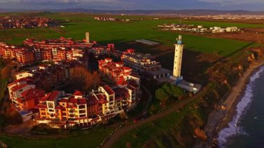 Yukarıdan, kırmızı çatılı binaları olan büyüleyici bir sahil köyü yemyeşil bir manzaranın yanında otururken, gün batımında uzun bir deniz feneri gururla kıyıda durur..