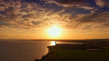 Güneş ufukta batarken turuncu ve sarı renkli canlı renkler gökyüzünü ve sakin suları aydınlatıyor. Huzurlu kıyı şeridi tefekkür ve akşam yürüyüşleri için mükemmel bir zemin sunuyor..