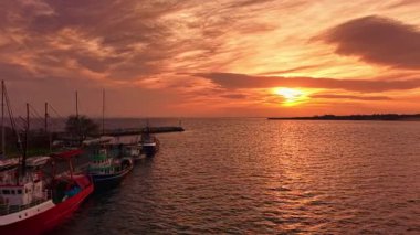 Güneş suyun üzerinde batarken altın renkler gökyüzünü dolduruyor. Balıkçı tekneleri limana demirlemiş, sakin bir manzara yaratıyorlar. Gece yaklaşırken yumuşak dalgalar gövdelere çarpar..