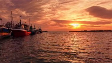 Balıkçı tekneleriyle dolu bir limanın üstündeki renkli günbatımı gökyüzünü boyuyor. Su ılık renklerle parıldıyor, gündüz geceye geçerken huzurlu bir atmosfer yaratıyor..
