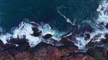 Kristal berrak dalgalar kıyı şeridinin karanlık kayalarına karşı çarpıcı bir kontrastı oluşturarak, öğleden sonra güneş ışığıyla aydınlatılıyor. Sahne doğanın güzelliğini ve gücünü yakalar..