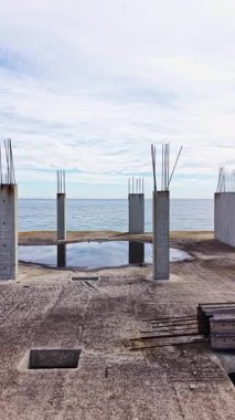 Beton sütunlar, okyanus kıyısındaki ıssız bir inşaat sahasında, bulutlar tepemizde süzülürken sakin suları yansıtarak, doğa ve insan gayreti arasında bir zıtlık yaratır..