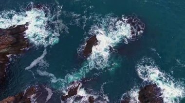 Dalgalar suyun kenarındaki kayalık oluşumlara karşı kabararak beyaz köpük oluşturur ve parlak mavi bir gökyüzü altında su sıçratır. Bu da doğanın dinamik güzelliğini cennet sahnesinde gösterir..