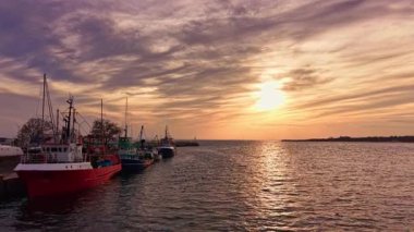 Parlak kırmızı ve yeşil balıkçı tekneleri güneş batarken parlayan sularla tezat oluşturur. Huzurlu bir marina sahnesinde gökyüzüne sıcak renkler saçar..