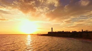 Güneş ufukta batarken, ılık altın renkler sakin okyanustan yansıyor. Arka planda bir deniz feneri duruyor. Huzurlu bir sahili çerçeveliyor..