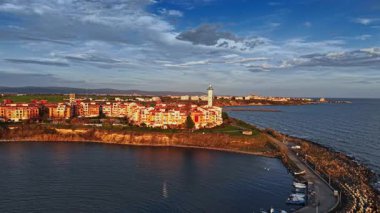 Gün batımında, kıyıda renkli binalar ve uzun bir deniz feneri bulunan bir sahil köyü üzerinde altın bir parıltı yayılıyor. Sakin sular güzel gökyüzünü yansıtır..