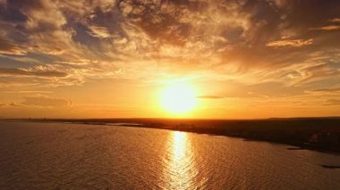 Canlı bir günbatımının nefes kesici manzarası suya altın renkler döküyor, uzak binaların siluetlerini parlaklaştırıyor. Sakin dalgalar renkli gökyüzünü yansıtır.