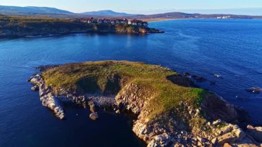 Altın güneş ışığı sakin bir kıyı şeridini aydınlatıyor, çimlerle kaplı kayalık bir çıkıntıyı aydınlatıyor, ışıl ışıl mavi sular ve uzak tepeler tarafından huzurlu bir geri çekilme sunuyor..