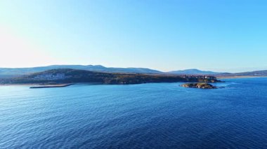 Sakin bir sahil manzarası sergileniyor, sakin sular parlak mavi gökyüzünü yansıtıyor. Uzaktaki tepeler ufku sarıyor dalgalar usulca kıyıları selamlıyor, huzur fısıldıyor.