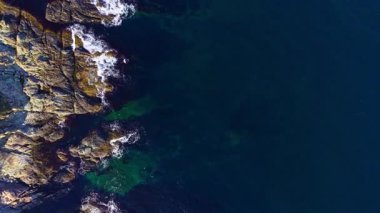 Dalgalar kıyı şeridine beyaz köpük dökerken, turkuaz sular pürüzlü kayalar arasında dans ediyor. Güneş ışığı, okyanus yüzeyinde parıldayan bir etki yaratır..