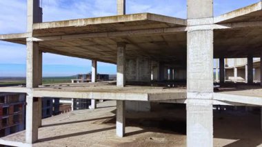 Büyük, bitmemiş bir yapı, parlak mavi gökyüzüne karşı yükselir, çıplak beton zemini ve çevresindeki geniş manzarayı gözler önüne serer, hırs ve terk edilme hakkında anlatılamamış hikayelere işaret eder..