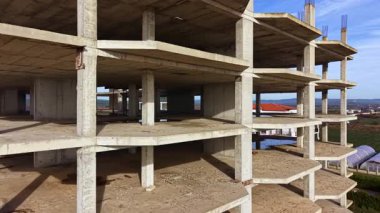 Bir inşaat sahasında, gökyüzüne doğru çıplak bir beton yapı yükseliyor, uzak tepelerle çevrili barışçıl bir manzaranın ortasında hala gelişmekte olan katlar sergileniyor..