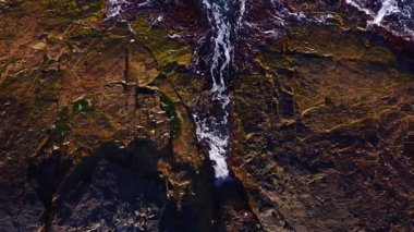 Güneş engebeli bir kıyı şeridinin üzerinde batıyor, desenli kayaların ve çarpan dalgaların üzerine sıcak renkler döküyor. Sahne, doğanın güzelliğini ve alacakaranlıktaki kara ve denizin uyumunu yakalıyor..