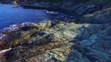 Engebeli kayalar kristal berraklığında mavi sulara karışıyor. Dalgalar parlak bir gökyüzünün altında, taşlı kıyıya doğru yavaşça süzülüyor. Doğanın güzelliği her ayrıntıda parlar..