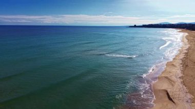 Altın kumlar, berrak bir gökyüzünün altında parlak mavi suların ufukla buluştuğu huzurlu bir kıyı şeridi boyunca uzanır. Yumuşak dalgalar bu sakin ortamda rahatlatıcı bir ritim yaratır..