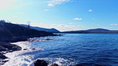 Dalgalar yavaşça kayalık kıyılara vururken, uzak tepeler canlı mavi gökyüzüne karşı yükselirken, gün ışığında doğal güzelliklerle dolu huzurlu bir sahil manzarası yaratıyor..