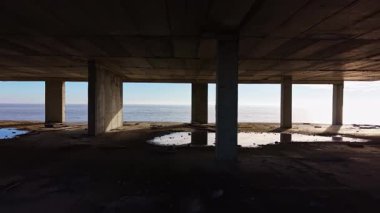 Güneş ışığı, ıssız bir binanın uzun beton sütunlarında sıcak bir parıltı saçar, sakin okyanus suları ve uzak ufuklar için sakin bir çerçeve oluşturur, nostalji ve sükunet hissi uyandırır..