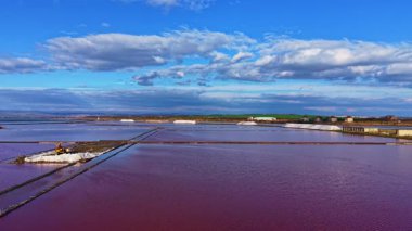 Parlak mavi bir gökyüzünün altında, sakin pembe tuz düzlükleri altın saat ışığında parıldarken, dağınık bulutlar durgun suların üzerinde tembelce sürükleniyor, pitoresk bir manzara yaratıyor..