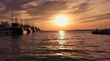 Balıkçı tekneleri rıhtımda sıralanırken altın güneş ışığı sakin sulardan yansıyor. Sıcak ve huzurlu bir gece, yaklaşan geceyi karşılayan sessiz bir limanda başlıyor..