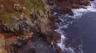 Güneş bu uzak kıyı sahnesinde batarken engebeli uçurumların tepesinde. Yumuşak dalgalar taşlı plajda süzülürken, desenli kayalar kıyı boyunca süzülüyor ve sakin bir atmosfer yaratıyor..