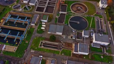 Hava görüntüsü, bir atık su arıtma tesisinin kapsamlı planını, sanayi bölgesindeki dairesel ve dikdörtgen tankları, boruları ve yakınlardaki binaları görüntüler..