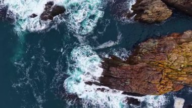 Dalgalar kıyı boyunca uzanan sivri kayalarla çarpışır, parlak güneş ışığı altında parlar. Sahne, okyanusun dinamik gücünü sakin bir ortamda yakalıyor..