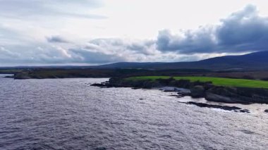 Kara bulutlar yukarıda toplanırken dalgalar engebeli kıyı şeridine hafifçe çarpıyor. Su kenarında yemyeşil tepeler yükselir ve günün kenarında çarpıcı bir kontrastlık yaratır..