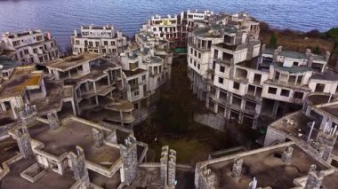 Doğa terk edilmiş deniz kenarındaki binaları geri alırken gölgeler ufalanan mimariye yayılıyor. Durgun su, sönen ışığı yansıtır. Ürkütücü ve büyüleyici bir sahne yaratır..