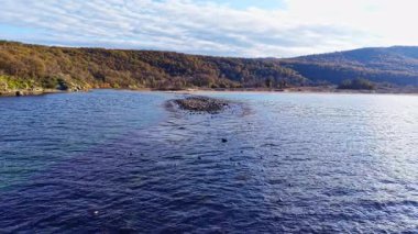 Sakin sular etrafındaki manzarayı yansıtır, tepeler yavaşça arka planda yuvarlanır. Işıldayan yüzeyden küçük bir kayalık ada belirir, tefekküre yol açar..
